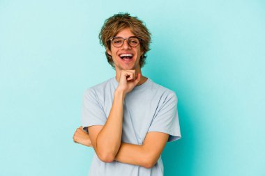 Mavi arka planda soyutlanmış, mutlu ve kendinden emin gülümseyen, elleriyle çenesine dokunan beyaz bir genç..