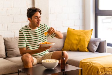 Genç melez adam kanepede oturmuş patlamış mısır yiyor.