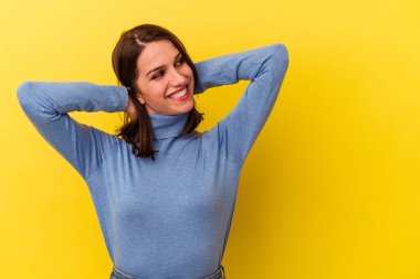 Sarı arka planda izole edilmiş genç beyaz bir kadın kendini güvende hissediyor, elleri başının arkasında..