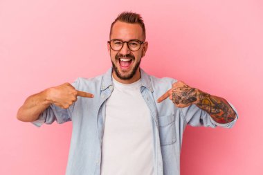 Pembe arka planda izole edilmiş beyaz dövmeli genç bir adam parmaklarıyla işaret ederek şaşırdı ve gülümsedi..