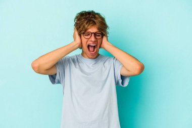 Makyajlı genç beyaz adam, mavi arka planda izole edilmiş kulakları kulaklarıyla kapatıyor ve çok ses duymamaya çalışıyor..