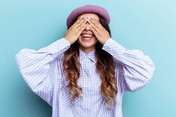 Mavi arka planda izole edilmiş genç Meksikalı kadın elleriyle gözlerini kapatıyor, gülümseyerek bir sürpriz bekliyor..