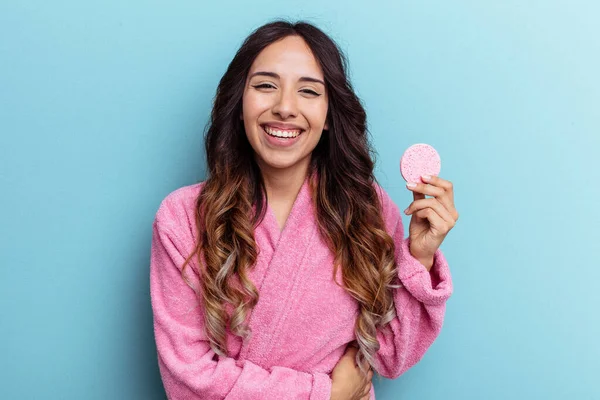Young mexican woman wearing a bathrobe holding a make-up remover sponge isolated on blue background laughing and having fun.
