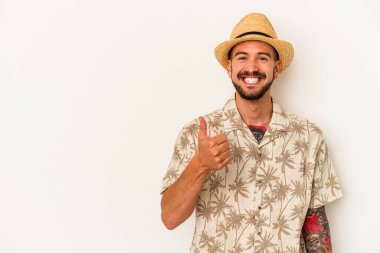 Beyaz arka planda izole edilmiş yaz kıyafetleri giyen dövmeleri olan genç beyaz bir adam gülümsüyor ve başparmağını kaldırıyor.