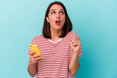 Elinde cep telefonu olan beyaz bir kadın mavi arka planda izole edilmiş ve ağzı açık bir şekilde üst tarafı işaret ediyor..