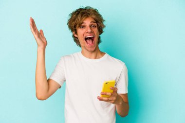 Makyajlı, cep telefonlu genç beyaz adam mavi arka planda izole edilmiş. Hoş bir sürpriz bekliyor, heyecanlı ve el kaldırıyor..