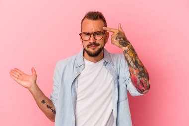 Pembe arka planda izole edilmiş dövmeleri olan genç beyaz bir adam elinde bir ürün tutuyor..