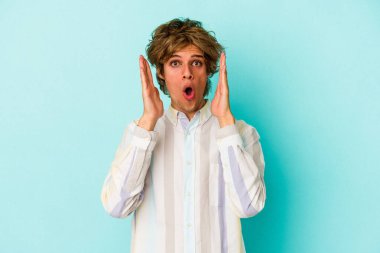 Makyajlı genç beyaz adam mavi arka planda izole edilmiş, şaşırmış ve şok olmuş..