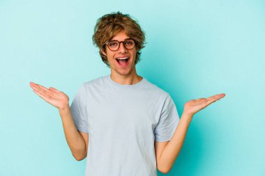 Mavi arka planda soyutlanmış beyaz makyajlı genç bir adam kollarıyla ölçeği yapıyor, mutlu ve kendinden emin hissediyor..