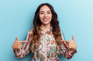 Mavi arka planda izole edilmiş genç Meksikalı kadın gömlek fotokopi alanını elle işaret ediyor, gururlu ve kendinden emin.