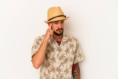 Yazlık giysiler giyen, beyaz arka planda izole edilmiş beyaz dövmeli genç beyaz bir adam şakağı parmağıyla işaret ediyor, düşünüyor ve bir göreve odaklanıyor..