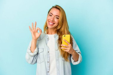 Elinde cep telefonu tutan beyaz kadın mavi arka planda izole edilmiş neşeli ve kendinden emin..