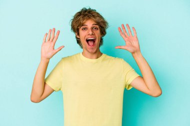 Mavi arka planda izole edilmiş beyaz tenli genç adam hoş bir sürpriz alıyor, heyecanlı ve el kaldırıyor..