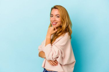 Beyaz kadın mavi arka planda izole edilmiş mutlu ve kendinden emin gülümsüyor, elleriyle çenesine dokunuyor..