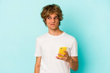 Makyajlı genç beyaz adam cep telefonunu mavi arka planda izole etmiş. Kafası karışmış ve kendinden emin değil..