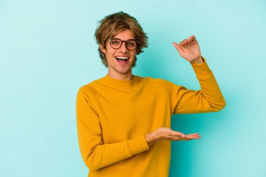 Mavi arka planda soyutlanmış beyaz makyajlı genç bir adam işaret parmaklarıyla küçük bir şey tutuyor, gülümsüyor ve kendinden emin..