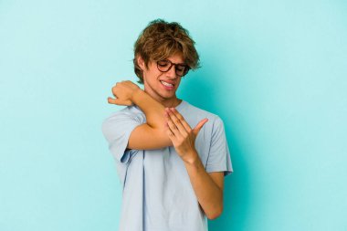 Makyajlı genç beyaz adam mavi arka planda izole edilmiş dirsek masajı yapıyor, kötü bir hareketin ardından acı çekiyor..