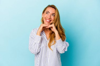 Mavi arka planda izole edilmiş beyaz kadın ellerini çenesinin altında tutuyor, mutlu bir şekilde yana bakıyor..