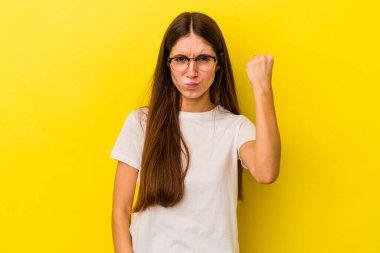 Sarı arka planda izole edilmiş genç beyaz kadın kameraya yumruğunu gösteriyor, agresif yüz ifadesi..
