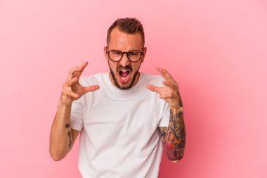 Pembe arka planda izole edilmiş dövmeleri olan genç beyaz bir adam öfkeyle bağırıyor..