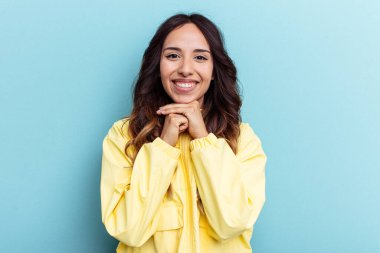 Mavi arka planda izole edilmiş genç Meksikalı kadın ellerini çenesinin altında tutuyor, mutlu bir şekilde yana bakıyor..