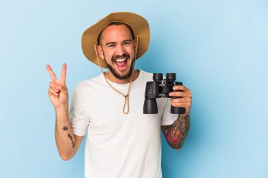 Mavi arka planda dürbün tutan beyaz dövmeli genç bir adam neşeli ve kaygısız bir barış sembolü gösteriyor..
