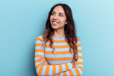 Mavi arka planda izole edilmiş genç Meksikalı kadın kollarını kavuşturarak gülümsüyor..