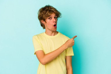 Makyajlı beyaz bir adam mavi arka planda izole edilmiş gülümseyen ve işaret eden boş bir alanda bir şey gösteriyor..