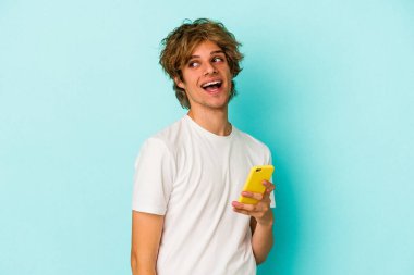 Makyajlı genç beyaz adam cep telefonunu mavi arka planda izole edip gülümsüyor, neşeli ve keyifli görünüyor..