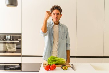 Genç melez adam öğle yemeği için salata hazırlıyor kameraya yumruğunu gösteriyor, agresif yüz ifadesi var..