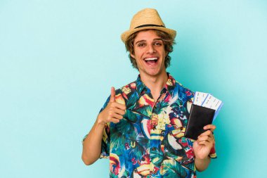 Makyajlı genç beyaz adam elinde pasaportuyla mavi arka planda izole edilmiş gülümsüyor ve başparmağını kaldırıyor.