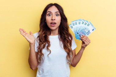 Sarı arka planda izole edilmiş banknotları tutan genç melez kadın şaşırdı ve şok oldu..