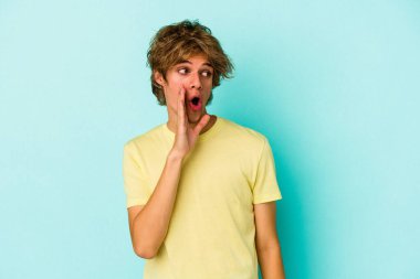Makyajlı genç beyaz adam mavi arka planda izole edilmiş. Gördüğü bir şey yüzünden şoka uğruyor..