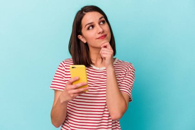 Mavi arka planda cep telefonu tutan beyaz bir kadın şüpheli ve şüpheci bir ifadeyle yana bakıyor..