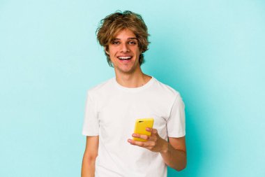 Makyajlı genç beyaz adam cep telefonuyla mavi arka planda izole edilmiş mutlu, gülümseyen ve neşeli..