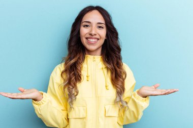 Mavi arka planda izole edilmiş genç Meksikalı kadın kollarıyla ölçeği yapıyor, mutlu ve kendinden emin hissediyor..