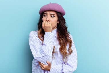 Mavi arka planda izole edilmiş genç Meksikalı kadın tırnaklarını yiyor, gergin ve çok endişeli..