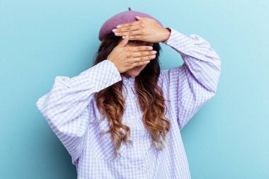 Mavi arka planda izole edilmiş genç Meksikalı kadın kameraya parmaklarıyla göz kırpıyor, yüzünü kapatmaktan utanıyor..