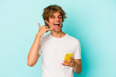 Makyajlı genç beyaz adam cep telefonuyla mavi arka planda izole edilmiş. Cep telefonu hareketini parmaklarıyla gösteriyor..