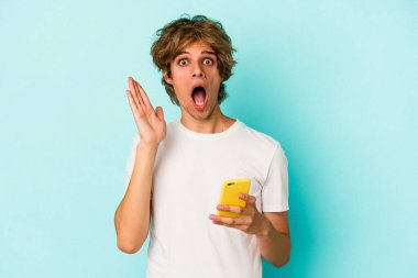 Makyajlı cep telefonlu genç beyaz adam mavi arka planda izole edilmiş, şaşırmış ve şok olmuş..