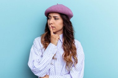 Mavi arka planda izole edilmiş genç Meksikalı kadın şüpheli ve şüpheci bir ifadeyle yana bakıyor..