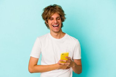 Makyajlı genç beyaz adam cep telefonunu mavi arka planda izole edip gülüyor ve eğleniyor..