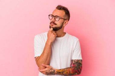Pembe arka planda izole edilmiş, şüpheli ve şüpheci bir ifadeyle yan bakan beyaz dövmeli genç bir adam..