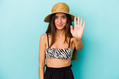 Genç beyaz kadın kumsala gidiyor. Mavi arka planda izole edilmiş bikini giyiyor. Beş numarayı parmaklarıyla gülümsüyor..