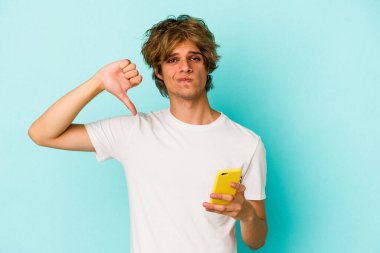 Makyajlı cep telefonlu genç beyaz adam mavi arka planda izole edilmiş. Sevimsiz bir hareket gösteriyor. Anlaşmazlık konsepti.