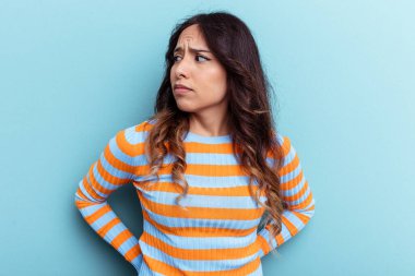 Mavi arka planda izole edilmiş genç Meksikalı kadın kafası karışmış, şüpheli ve güvensiz hissediyor..