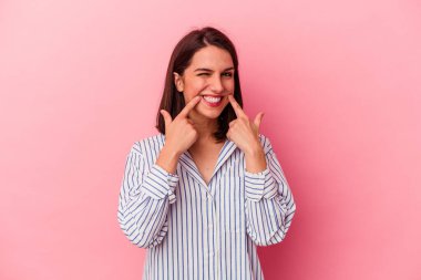 Pembe arka planda izole edilmiş genç beyaz kadın gülümsüyor, parmaklarını ağıza doğrultuyor..