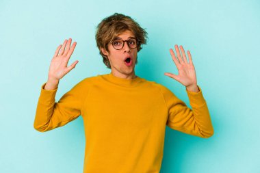 Makyajlı beyaz bir genç, mavi arka planda izole edilmiş. Yakın bir tehlike nedeniyle şok oluyor.