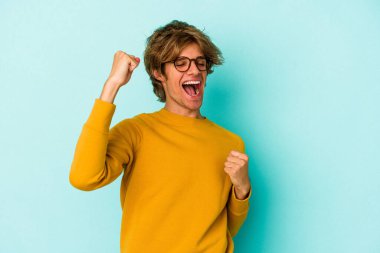 Makyajlı genç beyaz adam mavi arka planda izole edilmiş tasasız ve heyecanlı tezahüratlar yapıyor. Zafer konsepti.