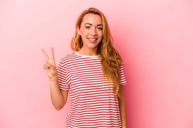 Pembe arka planda izole edilmiş beyaz sarışın kadın zafer işareti gösteriyor ve gülümsüyor..